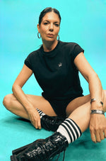 Woman sitting on a turquoise floor wearing a black outfit with striped socks and black boots.
